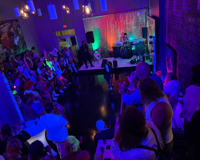 A large crowd gathers in a dimly lit venue, watching a performer on stage with colorful lights illuminating the space.