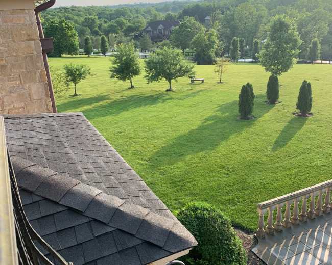 The image shows a view from a balcony overlooking a lush green lawn with several trees and a large field in the background.