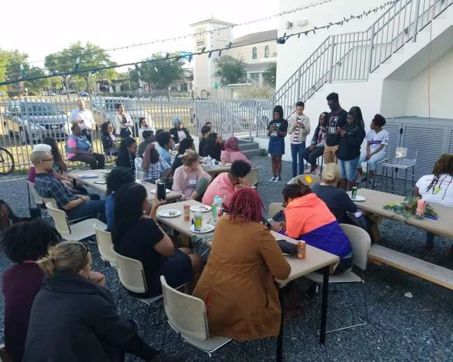 A group of people sits at tables outdoors, listening to speakers on a stage in front of a building with a staircase.