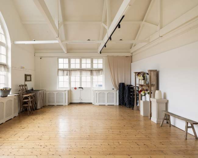 The image shows a spacious interior room with wooden flooring, large windows, and minimal furnishings, including a table, chairs, and cabinets along the walls.