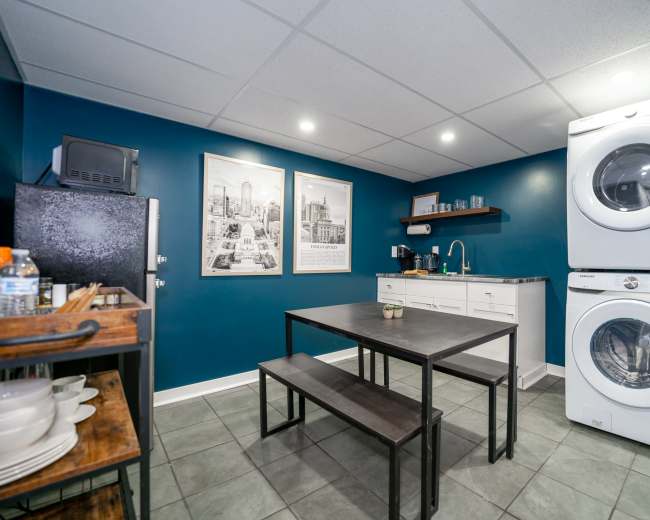 A modern kitchen space features a dark blue wall, a table with benches, and stacked washer and dryer units.