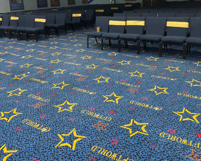 The image shows a spacious indoor area with rows of blue chairs arranged in a neat layout on a patterned carpet featuring stars and text.