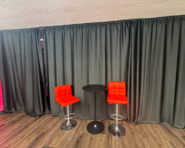 Two red bar stools are positioned next to a small black table in front of a black curtain backdrop.