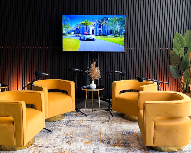 The scene features three yellow chairs arranged around a small table with a plant, and a large screen displaying an image of a car in front of a house on a black-paneled wall.