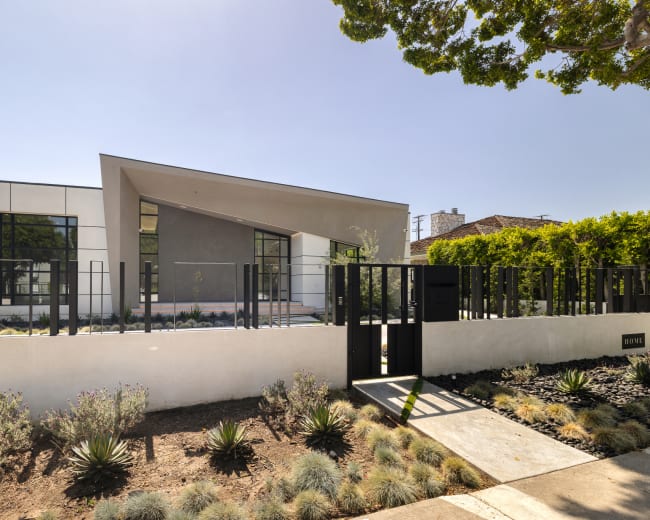 The image shows a modern house with a unique angular roofline, surrounded by a landscaped yard and enclosed by a stylish fence.