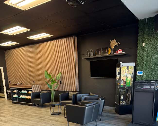 The image shows a modern waiting area with black chairs, a small table, and a green plant, set against a backdrop of wooden paneling and a television screen.