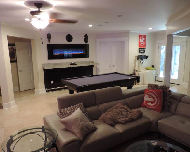 The image shows a spacious living area featuring a pool table, a sectional sofa, a modern fireplace, and access to an outdoor patio through glass doors.