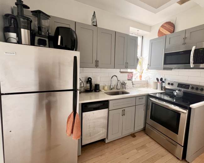 The image shows a modern kitchen with gray cabinets, stainless steel appliances, and a small sink area under a window.