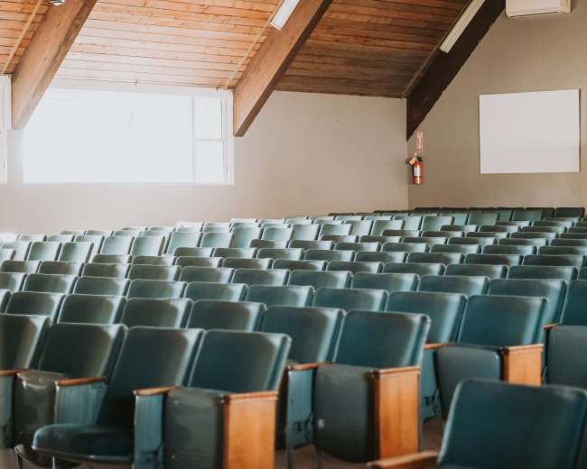 A nearly empty auditorium with rows of teal chairs arranged in a grid pattern beneath a wooden ceiling.
