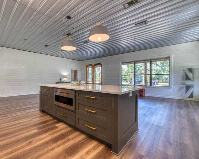 The image shows a modern kitchen with a central island, stainless steel appliances, and large windows offering a view of the outdoors.