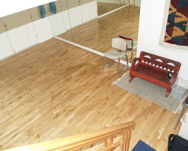 The image shows a wooden floor space with a chair, a red bench, and a large mirror reflecting the room.