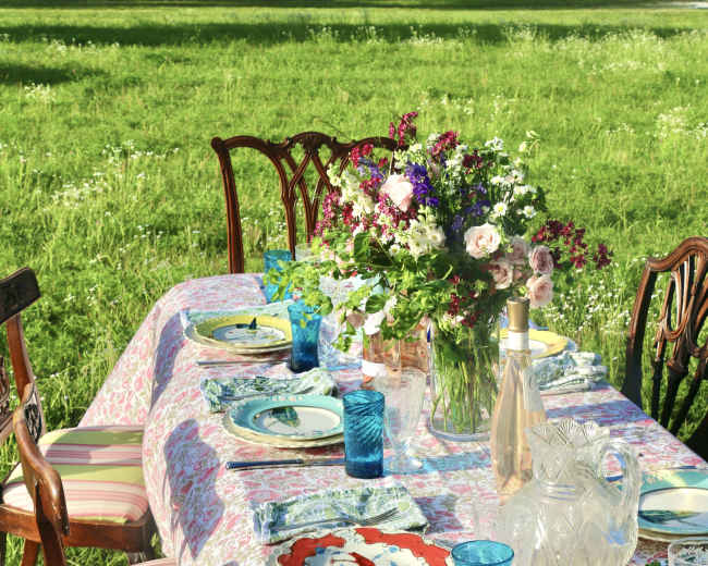 A table is set for a meal outdoors, adorned with colorful dishes, blue glasses, and a floral centerpiece, surrounded by a grassy area.