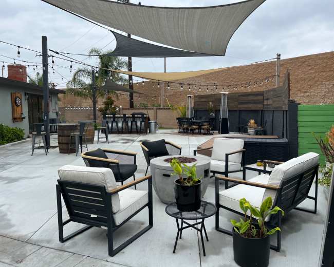 An outdoor patio area with modern seating, a fire pit, and shaded sections, surrounded by greenery and wooden fencing.