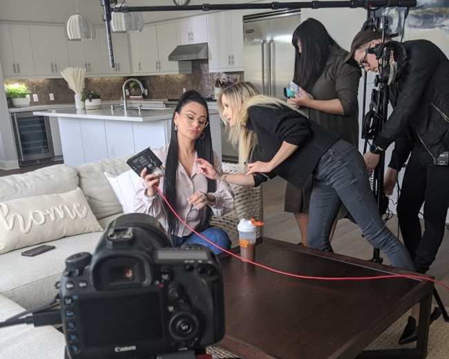 A woman is seated on a couch while a crew sets up a camera and equipment to film her in a modern living room.