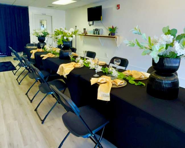 A decorated dining area features a long table set with floral centerpieces and dinnerware, surrounded by black chairs.