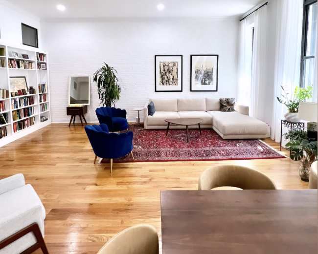 The image shows a modern living room with a white brick wall, a large beige sofa, a blue accent chair, a wooden dining table, and bookshelves lined with books.
