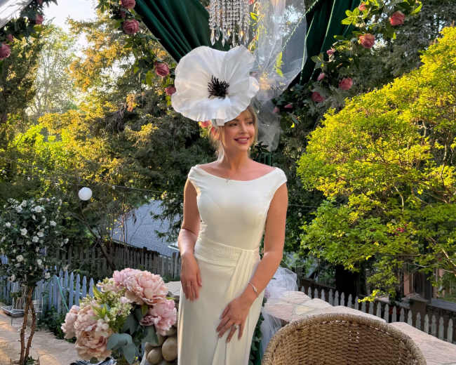A woman in a white dress stands in a garden setting, adorned with greenery and floral decorations, under a floral arrangement hanging above her.