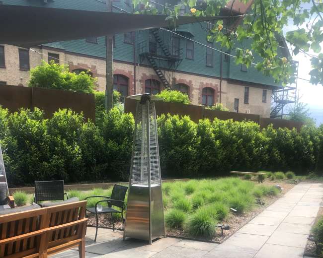 A landscaped outdoor space featuring a stone pathway, grassy areas, and a patio heater, with a multi-story building in the background.