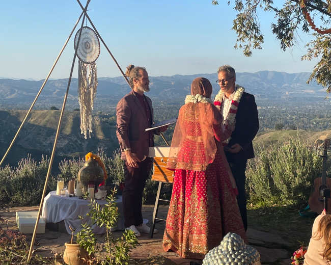A wedding ceremony is taking place outdoors on a hillside with mountains in the background, featuring a couple standing at an altar adorned with decorative elements.