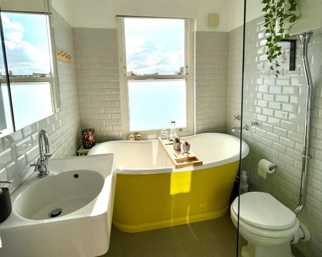 The bathroom features a freestanding yellow tub, a wall-mounted sink, and a glass shower enclosure with a large window allowing natural light.