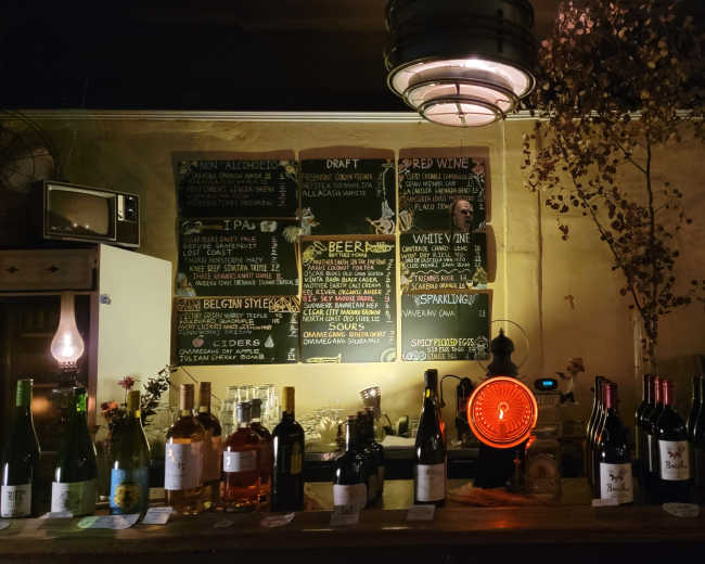 A dimly lit bar displays a collection of wine bottles and a chalkboard menu featuring various drinks.