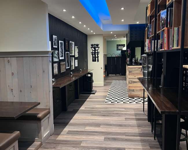 The image shows a modern cafe interior featuring wooden flooring, communal seating areas with tables along the walls, and decorative shelves filled with books and artwork.