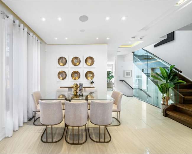 A modern dining area features a glass table surrounded by six upholstered chairs, with decorative wall plates and a staircase in the background.