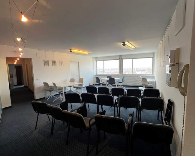 The image shows a spacious room arranged with rows of chairs facing a set of tables and large windows, illuminated by string lights.