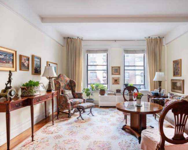 The living room features a round wooden table surrounded by chairs, with decorative art on the walls and large windows letting in natural light.