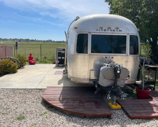 An Airstream trailer is parked on a concrete pad beside a red statue of a seated figure, with a grassy field in the background.