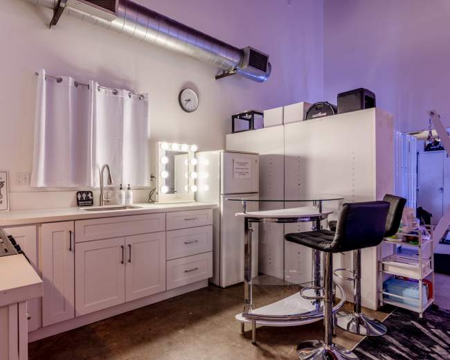 The image shows a modern kitchenette with white cabinetry, a sink, and a round bar table with a chair in an industrial-style setting.
