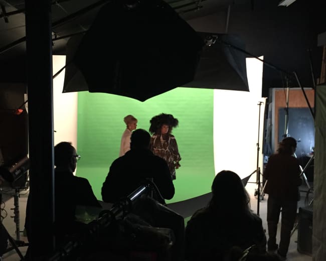 A group of people in a studio setting with a green screen, where two individuals are posing while others observe from the background.