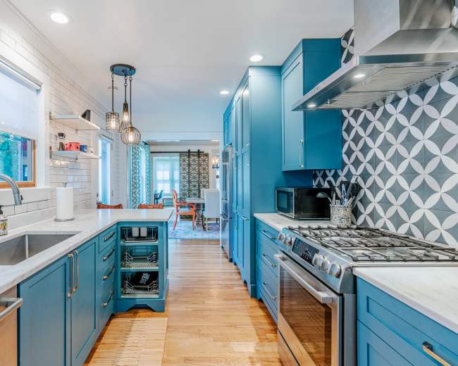 The image shows a modern kitchen with blue cabinets, a marble countertop, and stainless steel appliances, leading into a dining area with patterned curtains.