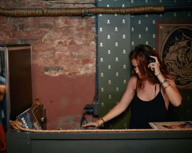 A DJ stands behind a mixer in a dimly lit room with exposed brick walls.