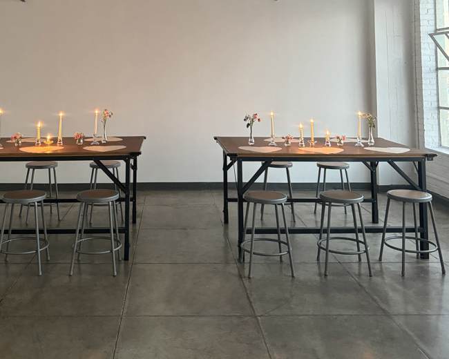 The image shows two tables arranged in a spacious room, each adorned with candles and small flower arrangements, surrounded by stools.