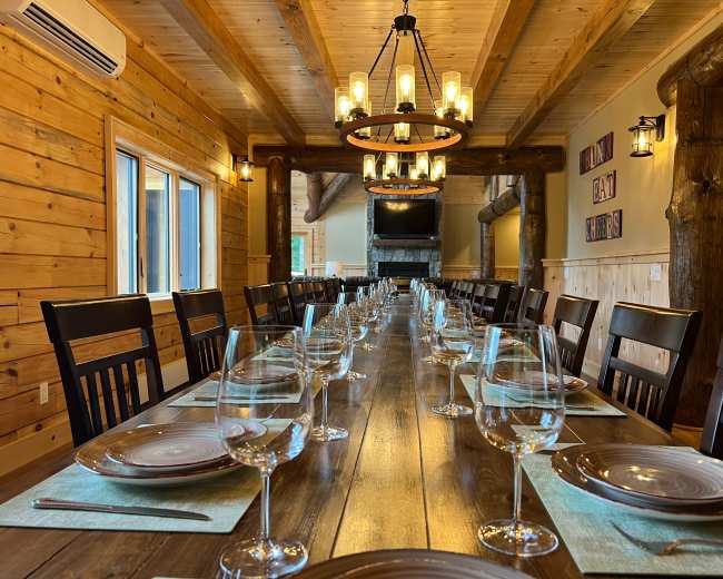 A long wooden dining table is set with plates and wine glasses in a rustic room with exposed wooden beams and a large chandelier.