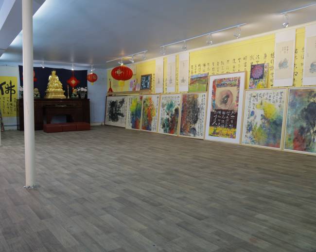 The image shows a spacious art gallery displaying framed colorful paintings along the walls, with a small altar and decorative elements at the back.