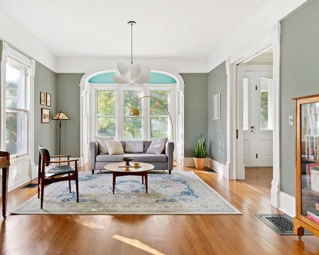 The image shows a well-lit living room featuring a gray sofa, a round coffee table, a piano, and large windows with green accents.