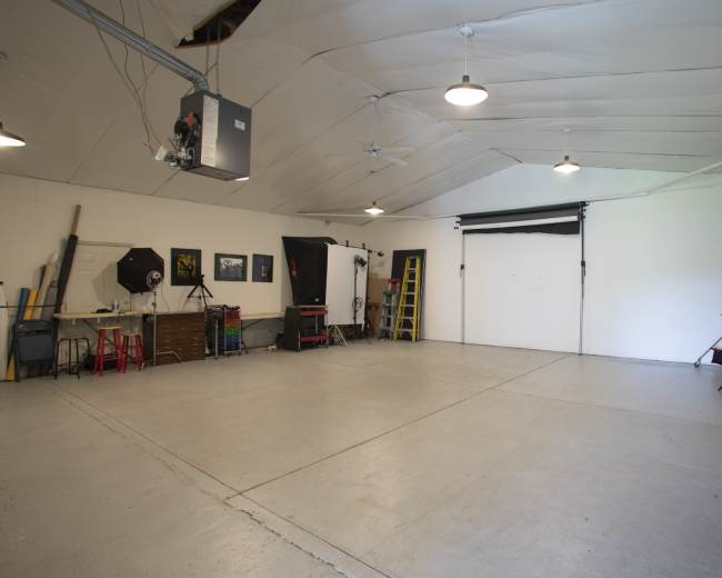 A spacious studio with white walls, a concrete floor, and photography equipment including lights, backdrops, and props organized along one side.
