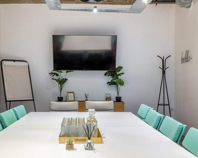 A modern meeting room features a large white table, green chairs, a television mounted on the wall, and potted plants.