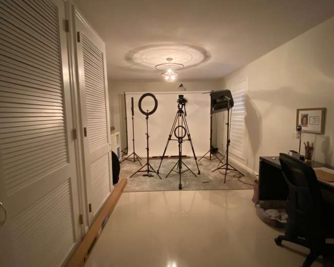 A well-lit indoor studio setup with a camera on a tripod, lighting equipment, and a desk in a corner.
