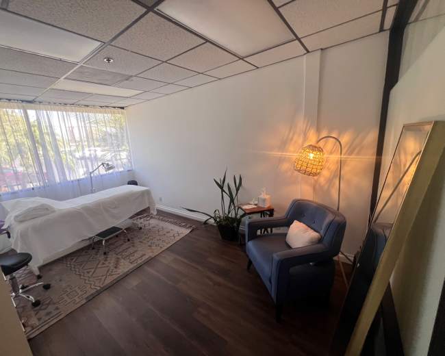 The image shows a bright and spacious therapy room with a treatment table, a chair, and a large window allowing natural light to enter.