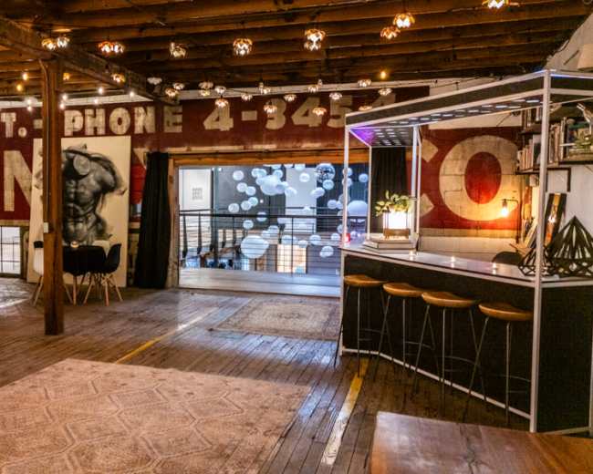 The image shows an interior space with exposed wooden beams, vintage advertising murals on the walls, and a bar area with modern furniture.