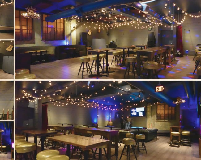 The image shows a spacious, dimly lit room designed for events, featuring wooden tables, bar stools, strings of decorative lights, and a stage area with sound equipment.