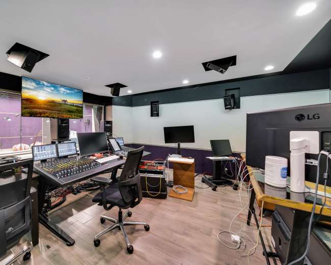 The image shows a modern audio recording studio equipped with a mixing console, multiple monitors, and various audio equipment in a well-lit environment.
