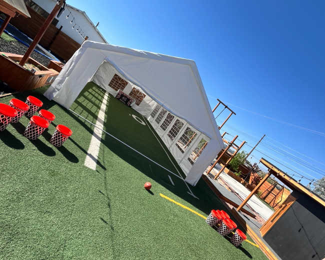 The image shows a white tent set up over a green turf field with red plastic containers arranged on one side.