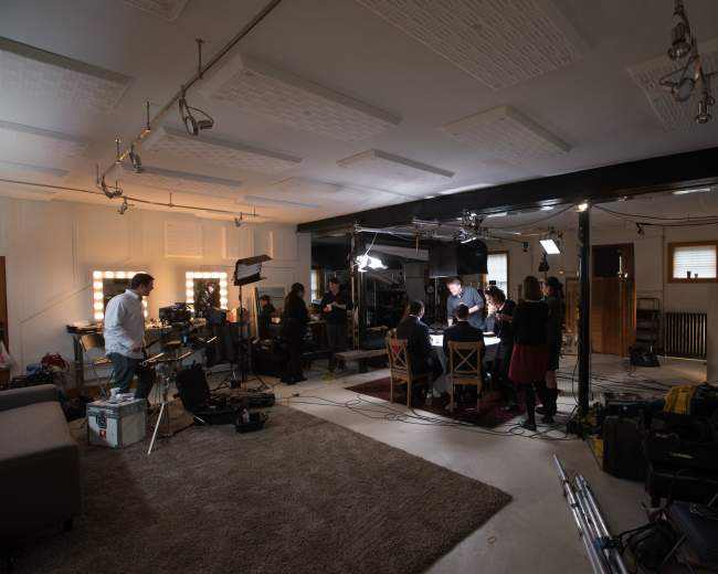 A video production studio features multiple crew members setting up equipment around a table for a shoot.