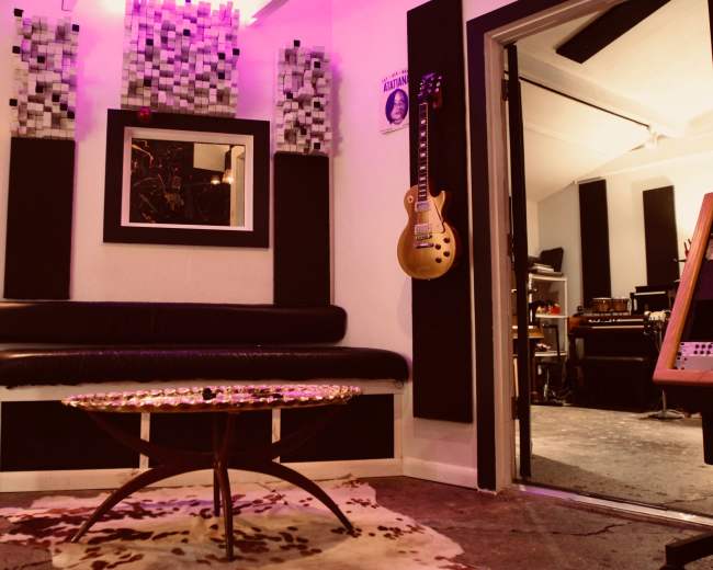 A music studio with a black couch, a circular coffee table, soundproof panels on the walls, and a guitar hanging nearby.