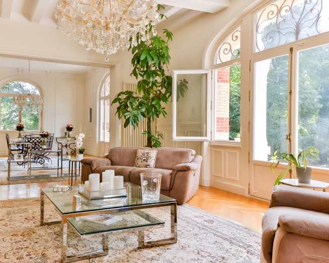 The image shows a bright living room featuring two brown sofas, a glass coffee table, a large chandelier, and an adjacent dining area with a table and chairs.
