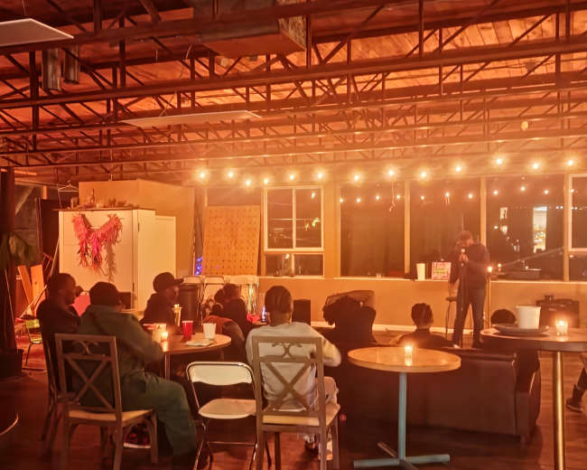 A small audience sits in a dimly lit venue, watching a performer on stage while surrounded by tables and string lights.